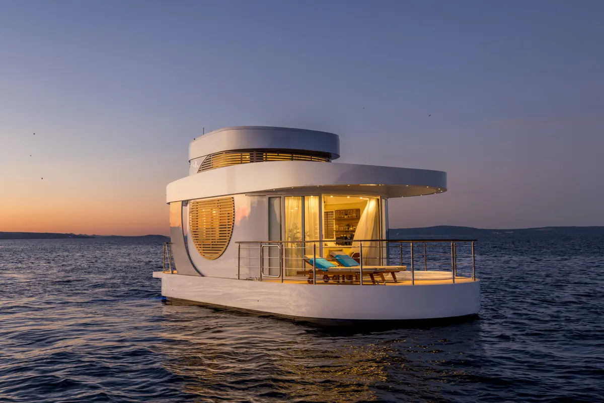 Amor from the side at dusk, the deck and interior lit, the last light on the horizon